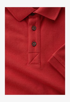 Tela de polo roja con tres botones marrones, cuello y manga corta con puño acanalado visible.