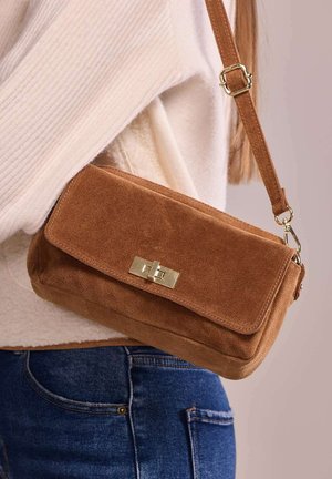 Sac bandoulière en daim marron avec fermeture dorée porté sur un pull crème et un jean bleu sur fond neutre.