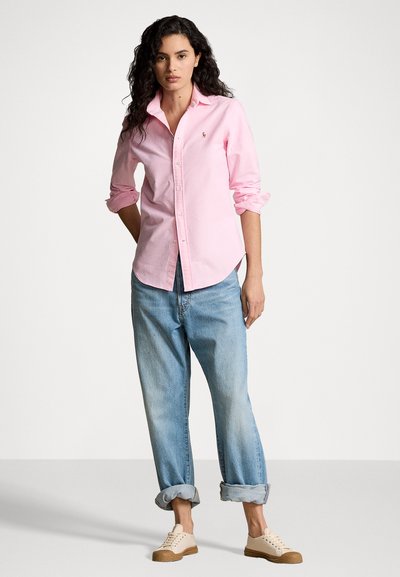 Helles rosa Hemd mit umgeschlagenen Ärmeln, kombiniert mit lockeren blauen Jeans und beigen Sneakers. Dezentes Logo auf dem Hemd.