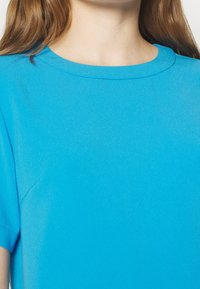 Modrý top s krátkým rukávem s kulatým výstřihem a hladkou texturou. Materiál vypadá lehce a je mírně lesklý.