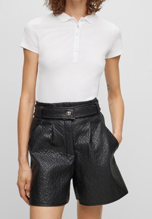 Kobieta ubrana w biały krótki rękaw polo oraz czarne, wysokowalne, teksturowane skórzane szorty z przednimi kieszeniami i paskiem zapinanym na guzik.
