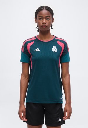 Kvinde med flettet hår iført en mørk teal og pink Real Madrid sportstrøje og sorte Adidas shorts, stående mod en ensfarvet baggrund.