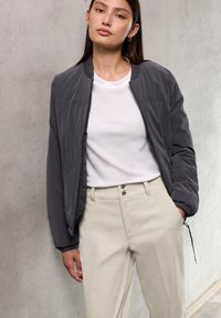 Jonge vrouw in een grijze bomberjack over een witte shirt en beige broek, staat tegen een lichtgrijze betonnen muur.