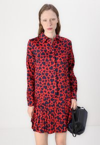 Abito rosso con un motivo a cuori blu navy, apertura frontale con bottoni e gonna plissettata. Abbinato a una borsa nera, caratterizzata da una texture liscia.
