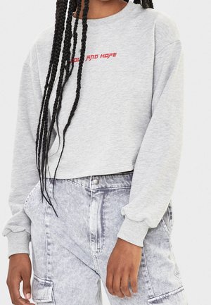 Personne portant un sweat court gris clair avec un texte rouge et un jean taille haute délavé à l'acide avec des poches latérales et de longues tresses.