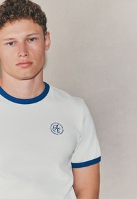 Kremowy t-shirt z krótkim rękawem, z niebieskim prążkowanym kołnierzykiem i mankietami. Na piersi znajduje się okrągłe granatowe logo z napisem. Wykonany z bawełny.