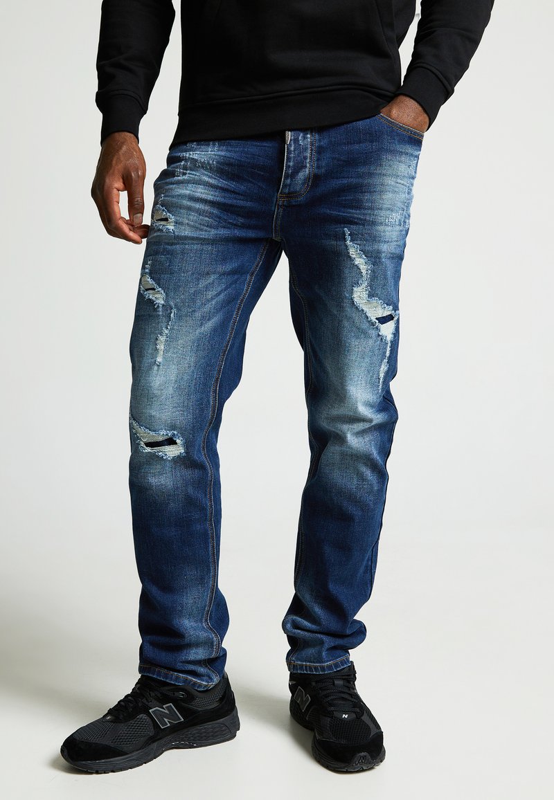 Vaqueros de denim azul con un ajuste entallado, que presentan desgarros desgastados y detalles desvaídos, combinados con zapatillas deportivas negras.