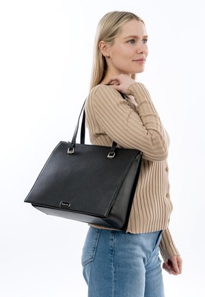 Vrouw met steil blond haar, gekleed in een beige geribbelde cardigan en blauwe spijkerbroek, met een grote zwarte leren shopper aan haar schouder.