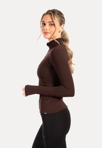 Haut marron à fermeture éclair avec manches longues, coupe ajustée et ouvertures pour les pouces aux poignets, assorti à un legging noir. Texture lisse, détails minimaux.