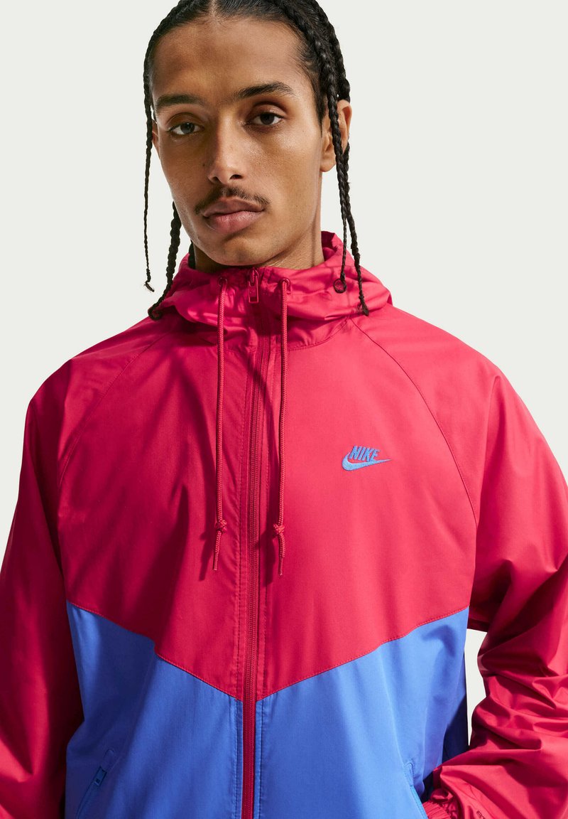 Man met gevlochten haar draagt een rode en blauwe Nike windjack met rits dicht, kijkend naar voren tegen een effen lichte achtergrond.