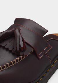 Σκούρο καφέ δερμάτινο παπούτσι τύπου loafer με φούντες, ορατά ράμματα και σόλα με κίτρινες λεπτομέρειες σε λευκό φόντο.