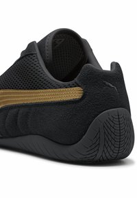 Scarpa sportiva nera con tomaia in suede testurizzato, striscia dorata e dettagli in rete. Presenta una suola in gomma con motivo di battistrada.