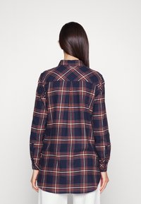 Navy plaid shirt with red and white check pattern, long sleeves, buttoned cuffs, and a classic collar. Features a curved hemline.