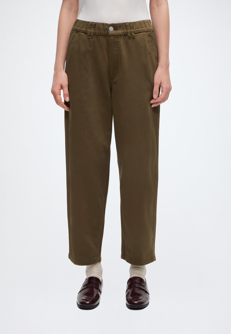 Άτομο που φορά ψηλόμεσο, άνετο χακί παντελόνι με κουμπιά στο μπροστινό μέρος, άσπρες κάλτσες και σκούρα καφέ δερμάτινα μοκασίνια, στέκεται μπροστά σε απλό λευκό φόντο.