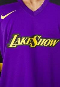 Purpursarkanais sporta krekls ar V-veida kaklu, uz kura ir uzraksts "LAKESHOW" izteiksmīgā dzeltenā fontā, akcentēts ar melniem plecu paneļiem un mazu dzeltenu logo.