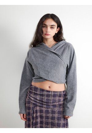 Młoda kobieta z długimi ciemnymi włosami, ubrana w szary krótki sweter w stylu kopertowym oraz wysoką fioletową kraciastą spódnicę na jednolitym tle.