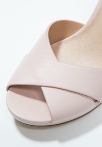 Gros plan d'une sandale rose pâle à bout ouvert avec des lanières croisées en cuir et une semelle intérieure légèrement texturée.
