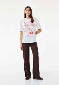 Witte T-shirt met een roze cocktailafbeelding en tekst. Gecombineerd met bruine geribbelde flared broeken. Model staat tegen een lichte achtergrond.