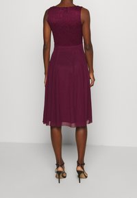 Burgundská krajková a látková šaty s vypasovaným korzetem a rozšířenou sukní, s přiléhavým pasem a délkou ke kolenům. Černé vysoké podpatky.