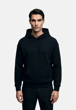 Hombre con sudadera negra con capucha y pantalones negros, de pie recto frente a un fondo claro y liso, con las manos relajadas a los lados.