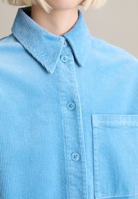 Lichtblauw corduroy overhemd met een button-down kraag, drie knopen en een borstzak met verticale ribbels.