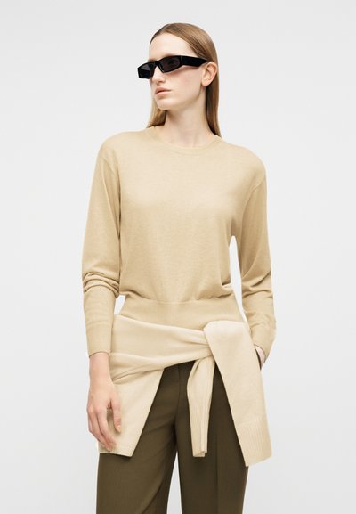 Femme portant des lunettes de soleil rectangulaires noires, un pull beige avec un autre pull assorti noué à la taille, et un pantalon vert olive sur fond blanc.