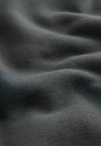 Schwarzer Baumwollstoff mit glatter Textur, der subtile Dehnbarkeit und eine matte Oberfläche aufweist. Die Fläche zeigt sanfte Falten und Wellen.