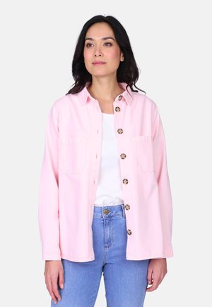 Femme portant une chemise rose clair boutonnée sur un haut blanc et un jean bleu, debout devant un fond clair uni.