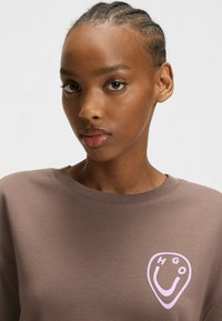 Sweatshirt marron avec un col rond et des poignets côtelés, arborant un logo rose clair en forme de bouclier sur la poitrine. Texture douce.