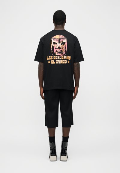 Άνδρας που φοράει μαύρο φαρδύ T-shirt με γραφικό από ροζ και πορτοκαλί μάσκα πάλης και το κείμενο «Les Benjamins El Gringo» στην πλάτη.