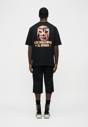 Άνδρας που φοράει μαύρο φαρδύ T-shirt με γραφικό από ροζ και πορτοκαλί μάσκα πάλης και το κείμενο «Les Benjamins El Gringo» στην πλάτη.