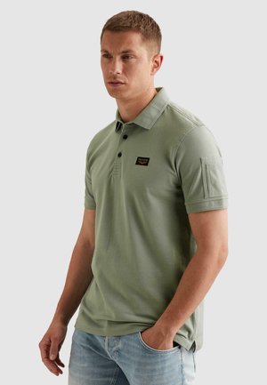 Mann trägt ein hellgrünes Polohemd mit kurzen Ärmeln und einem kleinen schwarzen Logopatch auf der Brust sowie hellblaue Jeans, steht mit einer Hand in der Tasche.