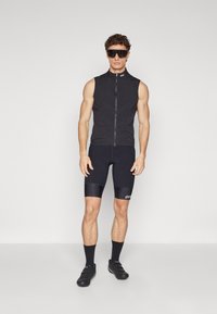 Gilet de cyclisme noir sans manches avec une fermeture éclair à l'avant, associé à un short noir ajusté avec une texture côtelée et un logo blanc. Chaussures de cyclisme noires.