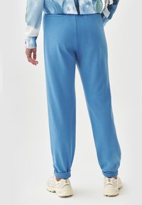 Blaue Sweatpants aus einem weichen Stoff, mit schmal zulaufendem Design und gerippten Knöcheln, kombiniert mit hellen Sportschuhen.