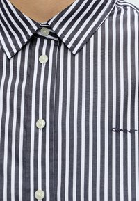 Chemise boutonnée rayée verticalement en noir et blanc avec col pointu et petit logo brodé "GANT" sur la poitrine.