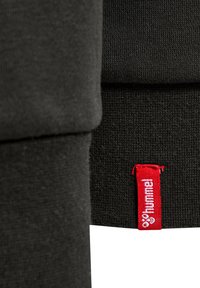 Svart textil med en slät yta, med en röd etikett som har texten "hummel". Designen har en ren, minimalistisk stil.