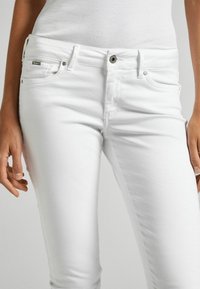 Vita denimjeans med en figurnära design, som har en dragkedja, framfickor och subtila sömnadsdetaljer som syns på tyget.