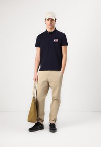 Marineblå polo med logo, khakifargede bukser, og svarte joggesko; holder en beige tote bag; kortermet, kroppsnær design, avslappet utseende.