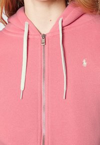Růžová mikina na zip vyrobená z měkké tkaniny, s předním zipem, bílými šňůrkami a malým logem na hrudi. Texturovaný povrch.