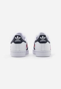 Sneakers bianchi con accenti blu navy, strisce rosse e superficie testurizzata. Presentano un tallone imbottito, suola piatta e logo Adidas sul retro.