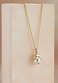 Kuldne kaelakee, millel on valge läikiv ripats, kaunistatud roheliste ja siniste elementidega, pehme roosa tausta ees.