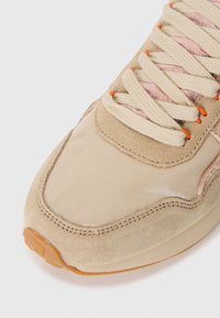 Baskets beige avec un embout en cuir lisse et brillant, une tige en daim, un système de laçage bien visible et des accents orange près des œillets.
