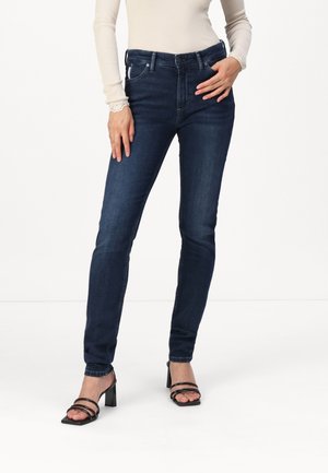 Jeans skinny bleu foncé en denim, avec une taille mi-haute, cinq poches et un léger décoloration sur le devant, assortis à des sandales à talons noires.