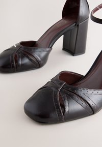 Talons pour femmes en cuir marron foncé avec bouts arrondis, couture perforée décorative et talons blocs épais sur une surface claire.