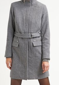 Šedý kabát s vlněným vzorem pepito, vysokým límcem, páskem v pase, dvěma předními kapsami a žebrovanými manžetami. Hladká textura z vlněné směsi.