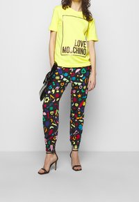 Żółta koszulka z napisem "LOVE MOSCHINO" w pogrubionych czarnych literach, zestawiona z czarnymi spodniami z kolorowymi nadrukami i dopasowanymi mankietami.