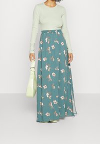 Maxihame teal dengan pola bunga, pinggang elastis, dan belahan samping. Dipasangkan dengan atas panjang lengan hijau muda dan tas bertekstur.