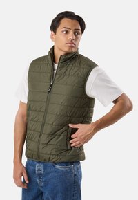 Jaunas vyras dėvi žalią pūstu liemenį be rankovių ant baltos marškinėlių ir mėlynų džinsų, žiūrintis į šoną, viena ranka kišenėje.