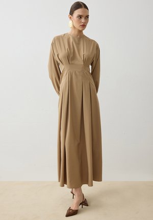 Kvinde iført en beige maxi-kjole med lange ærmer og høj talje med læg, brune spidse hæle og store guldbelagte øreringe, stående op ad en ensfarvet væg.