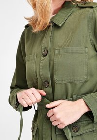 Olijfgroene jas van gestructureerde stof, met een kraag, een knoopsluiting aan de voorkant en twee borstzakken. Om de taille gestrikt met een riem.